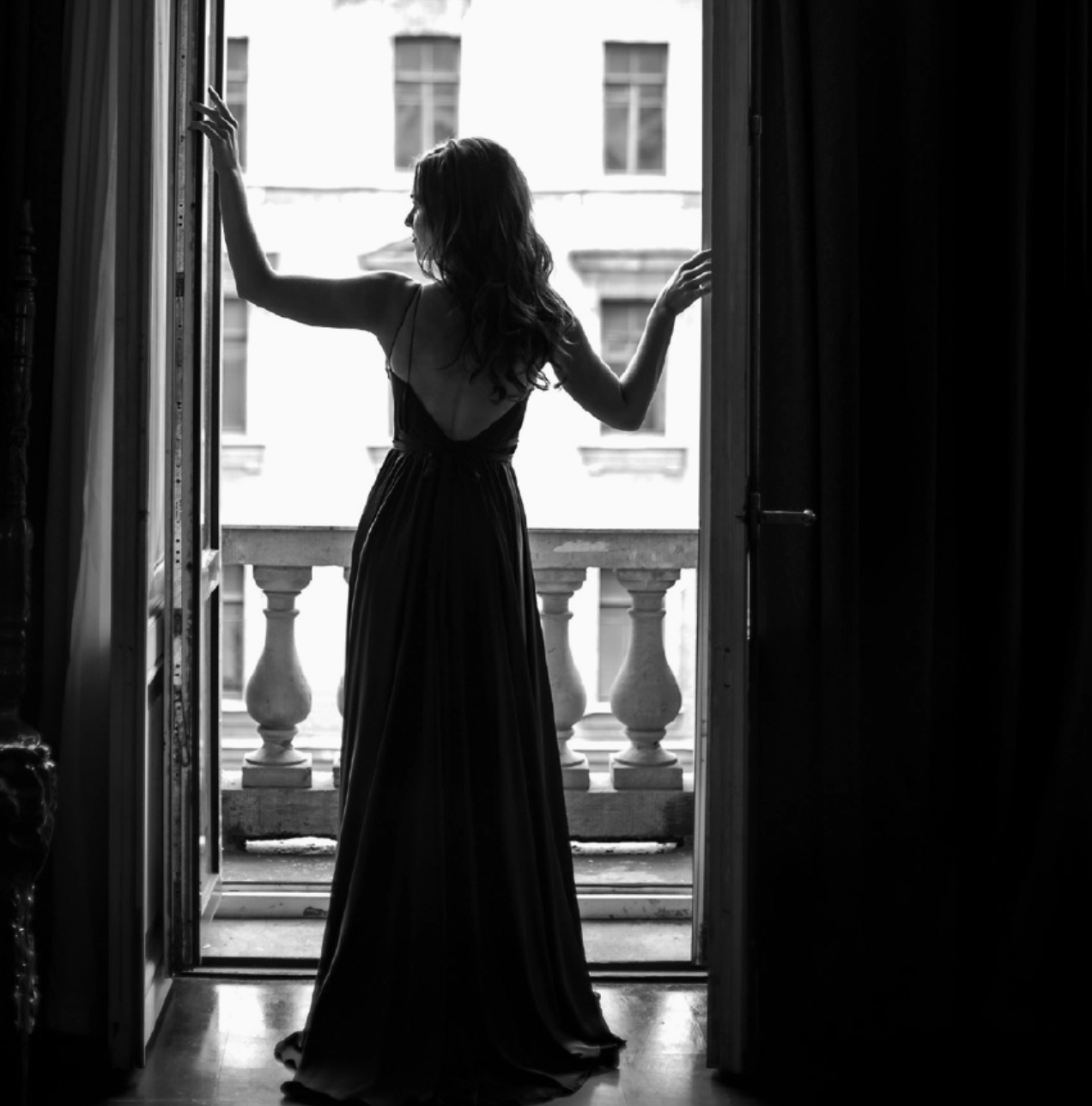 Woman in a long dress standing in a doorway with a balcony view.